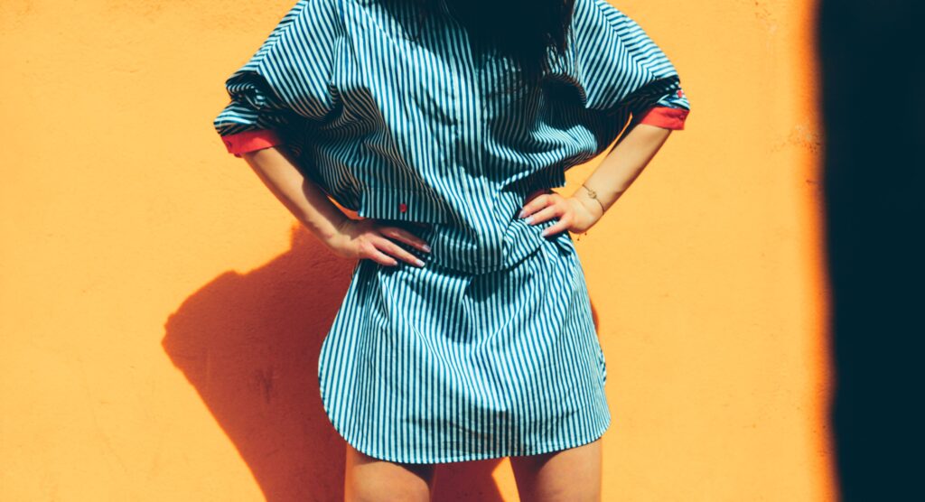 Fashionable woman posing in striped dress against a vibrant orange wall in sunny light.