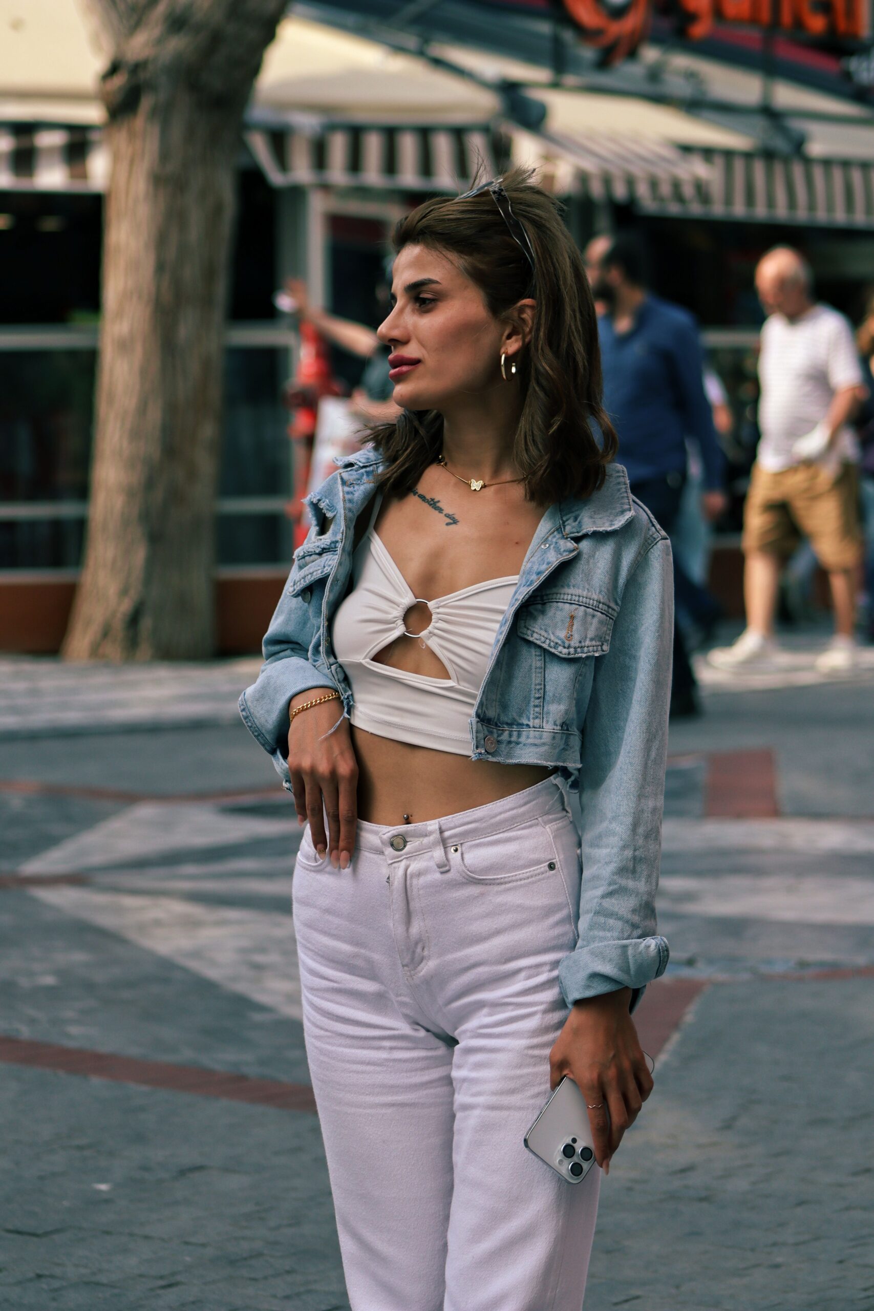 Stylish woman wears a jean jacket and holds a phone while standing outdoors on a city sidewalk.
