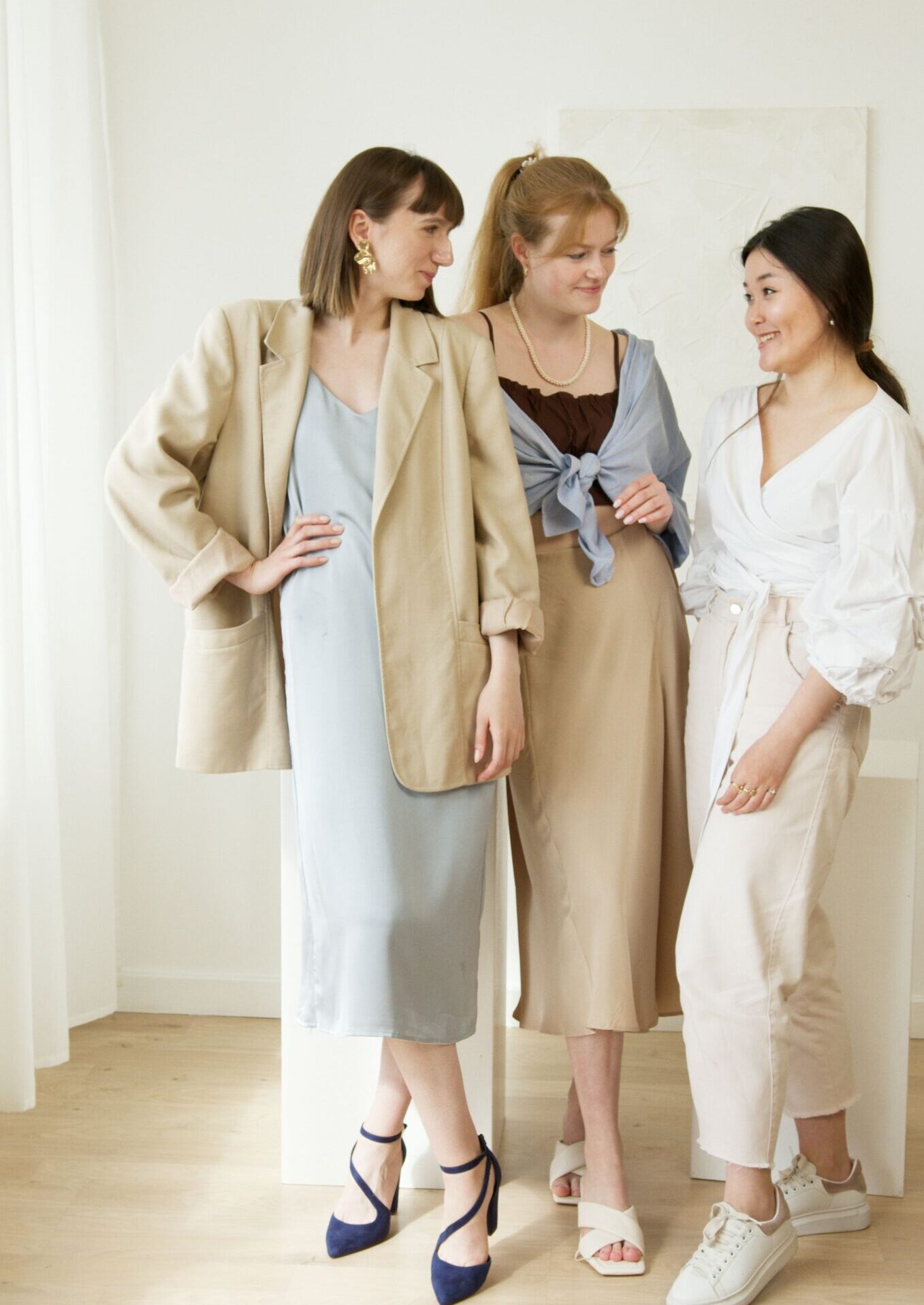 Three women in stylish outfits stand indoors, highlighting fashion and diverse beauty.