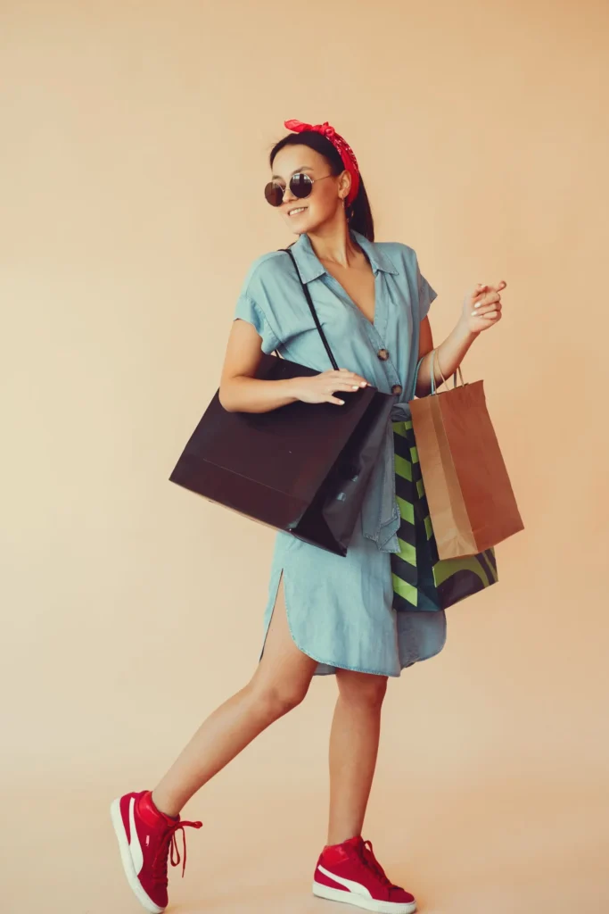 Woman wearing a light blue casual dresses with sunglasses and red sneakers, carrying shopping bag.