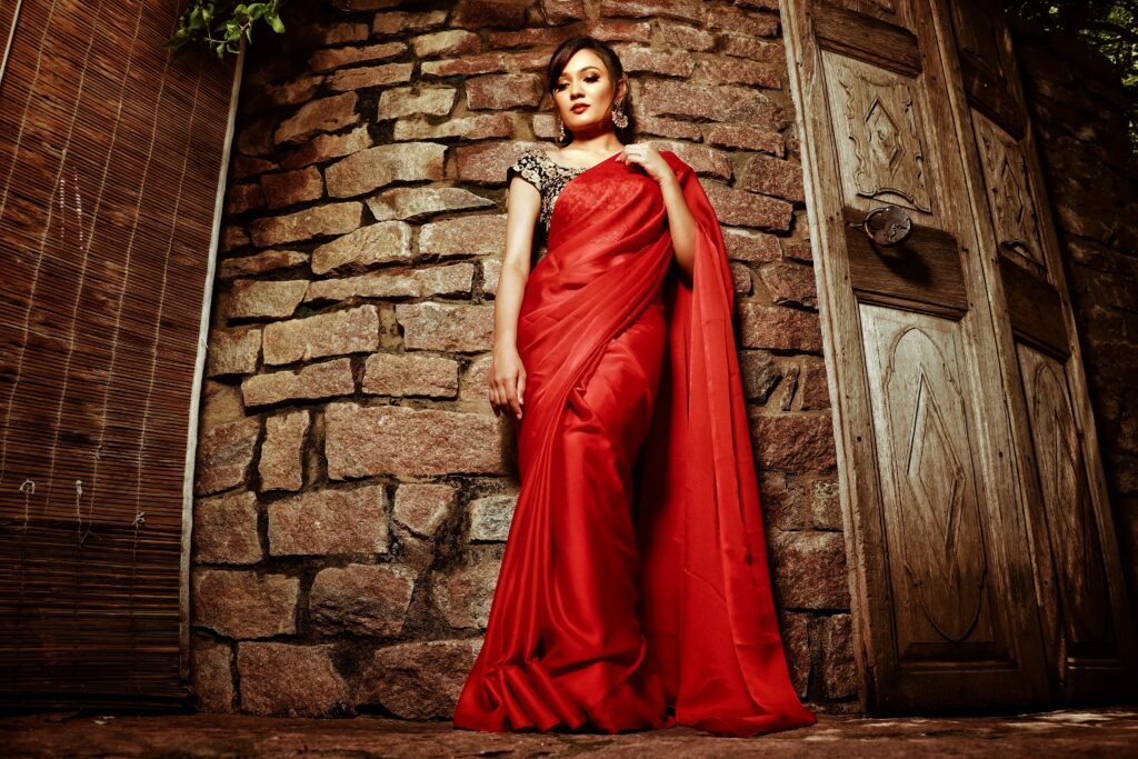 A woman in a red sari poses confidently against a stone wall with wooden doors.